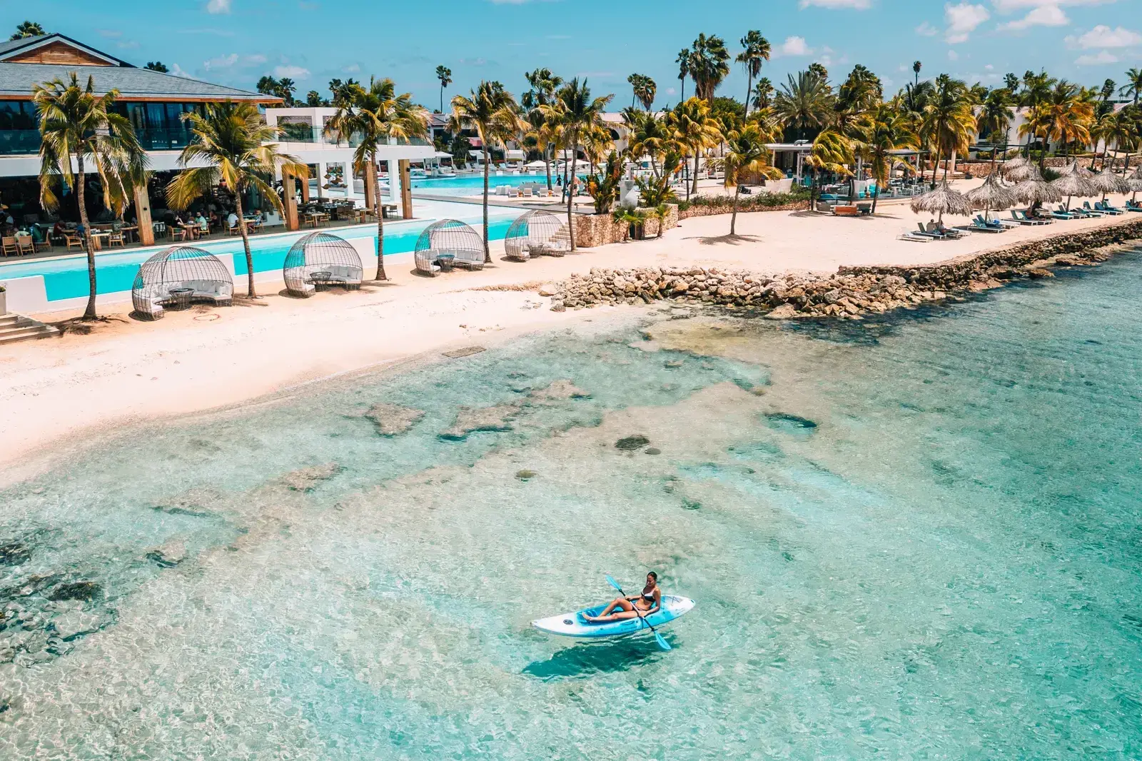 Luchtfoto van het strand en het heldere water van huisrif Eighteen Palm bij Van der Valk Bonaire, met een paddleboarder